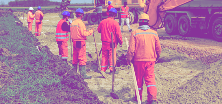 Plusieurs ouvriers creusent avec des pelles des tranchées sur un chantier avec des pelleteuses en arrière plan.