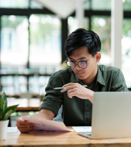 Un étudiant est assis, regarde un document de comptabilité avec un ordinateur posé en face de lui.