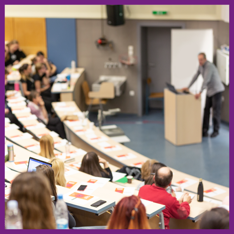 Plusieurs personnes sont assises dans un amphithéâtre et participent à un colloque animé par des professeurs.