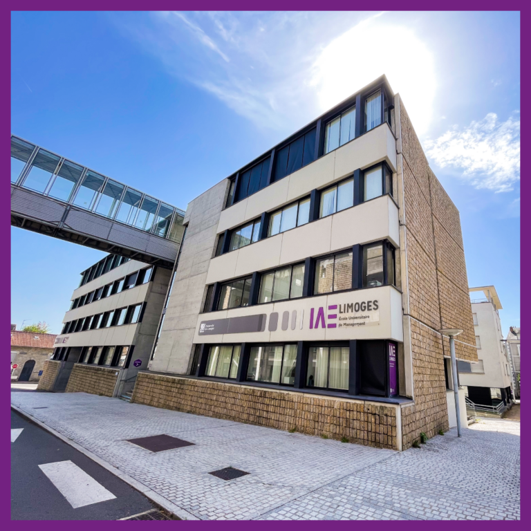 Bâtiment principal de l'IAE Limoges, École Universitaire de Management, sous un ciel bleu avec soleil éclatant en arrière-plan. On aperçoit également une passerelle vitrée reliant deux bâtiments.
