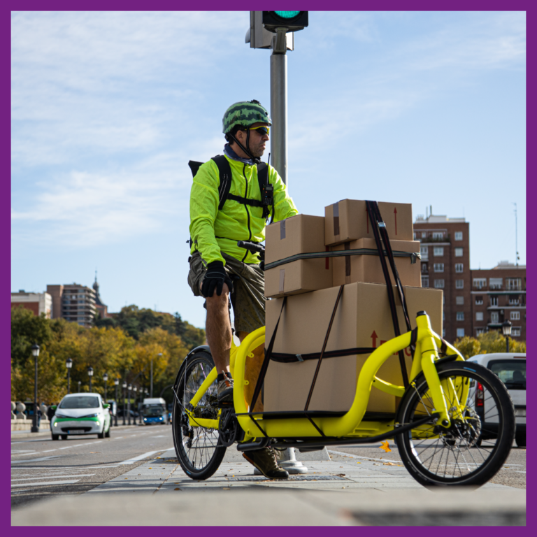 Un homme à vélo, avec plusieurs colis chargés sur son vélo cargo, est arrêté dans une grande ville au milieu d'un trottoir.