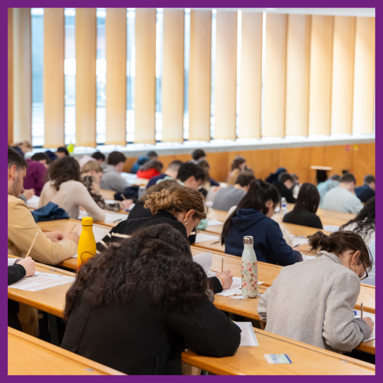 Étudiants dans un amphithéâtre en train de composer, concentrés sur leurs copies. Vue de dos avec plusieurs rangées de tables en bois, gourdes et fournitures scolaires posées sur les bureaux. L'éclairage naturel provient de grandes fenêtres partiellement couvertes par des stores.