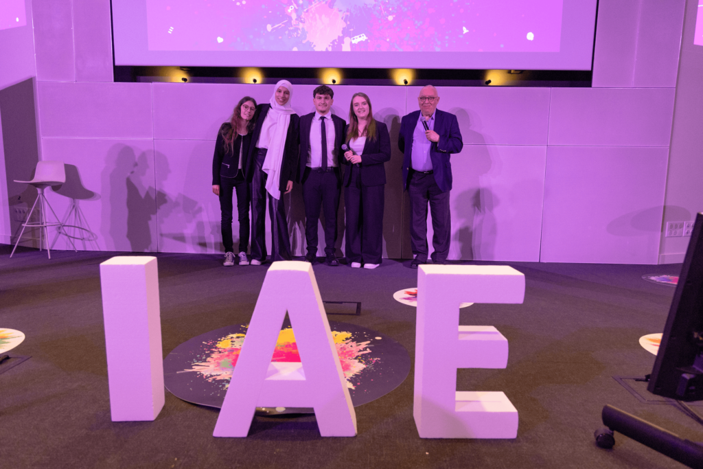 Remise de prix lors de la finale 2025 des Ateliers de l’innovation. Lauréats et membres du jury sont alignés sur scène, tenant diplômes et trophées, devant de grandes lettres formant « IAE ». L'événement met en lumière les projets innovants des étudiants.
