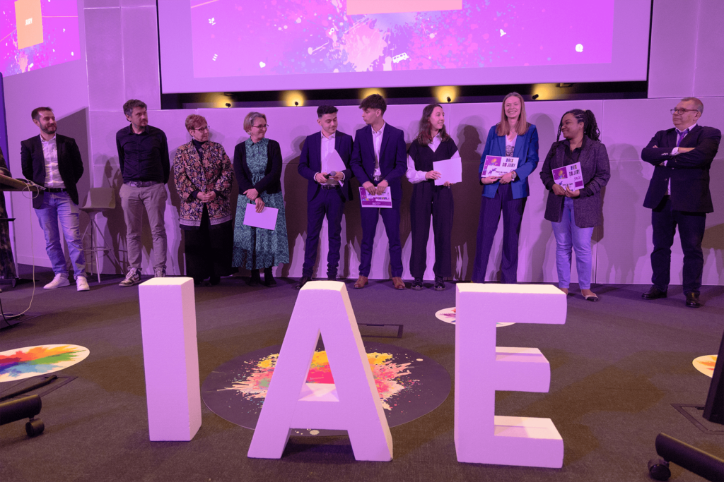 Remise de prix lors de la finale 2025 des Ateliers de l’innovation. Lauréats et membres du jury sont alignés sur scène, tenant diplômes et trophées, devant de grandes lettres formant « IAE ». L'événement met en lumière les projets innovants des étudiants.