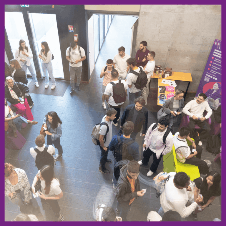 Vue en plongée d’un hall animé de l’IAE Limoges, rempli d’étudiants échangeant en petits groupes. Certains sont assis, d'autres debout, discutant ou consultant leur téléphone. À droite, un stand d'information présente des brochures et un kakémono violet sur le master management de l'innovation. L’ambiance est conviviale et dynamique.