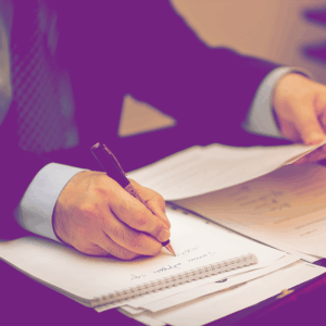 Un homme en costume rédige des notes à la main tout en consultant un document officiel, posé sur un bureau avec d'autres papiers administratifs.