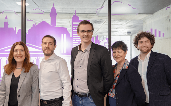 Cinq personnes posent ensemble devant une vitre ornée d’une illustration violette représentant une ville stylisée. Elles sont habillées en tenue professionnelle et sourient à l’appareil. L’arrière-plan laisse entrevoir un espace intérieur lumineux et moderne. De gauche à droite : Lisa Sharman, Directrice des Relations Internationales ; Hadrien Narbonne, Directeur de la Communication ; Gauthier Casteran, Directeur de l’IAE Limoges ; Martine Hlady-Rispal, Directrice Adjointe ; François Gravié-Plandé, Directeur des Études.